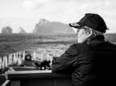 A conversation with the Commodore commanding SNMG1 Profile of a person wearing a cap and glasses, sitting on a bench by the sea with a blurred harbor in the background.