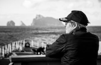 A conversation with the Commodore commanding SNMG1 Profile of a person wearing a cap and glasses, sitting on a bench by the sea with a blurred harbor in the background.