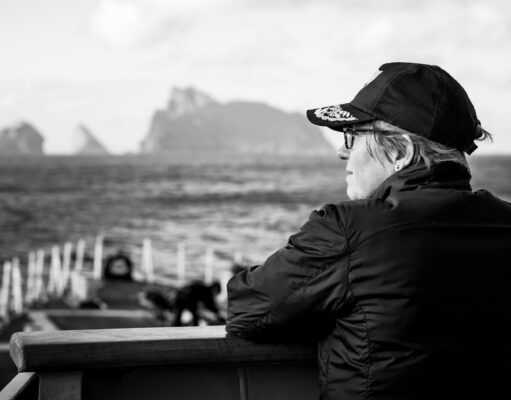 A conversation with the Commodore commanding SNMG1 Profile of a person wearing a cap and glasses, sitting on a bench by the sea with a blurred harbor in the background.