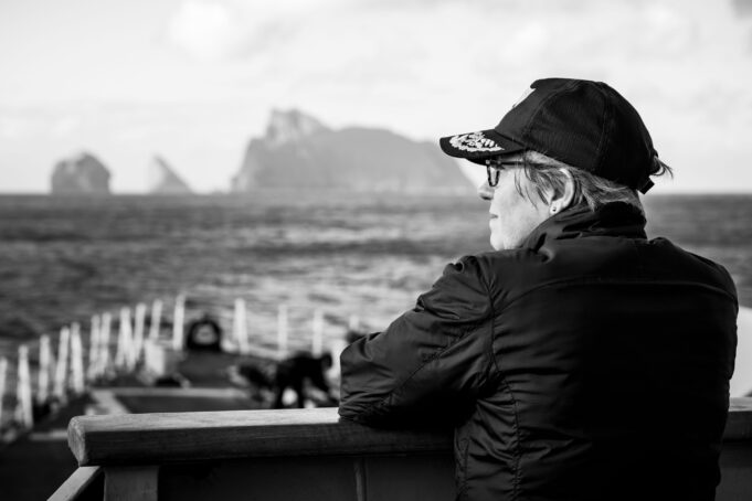 A conversation with the Commodore commanding SNMG1 Profile of a person wearing a cap and glasses, sitting on a bench by the sea with a blurred harbor in the background.