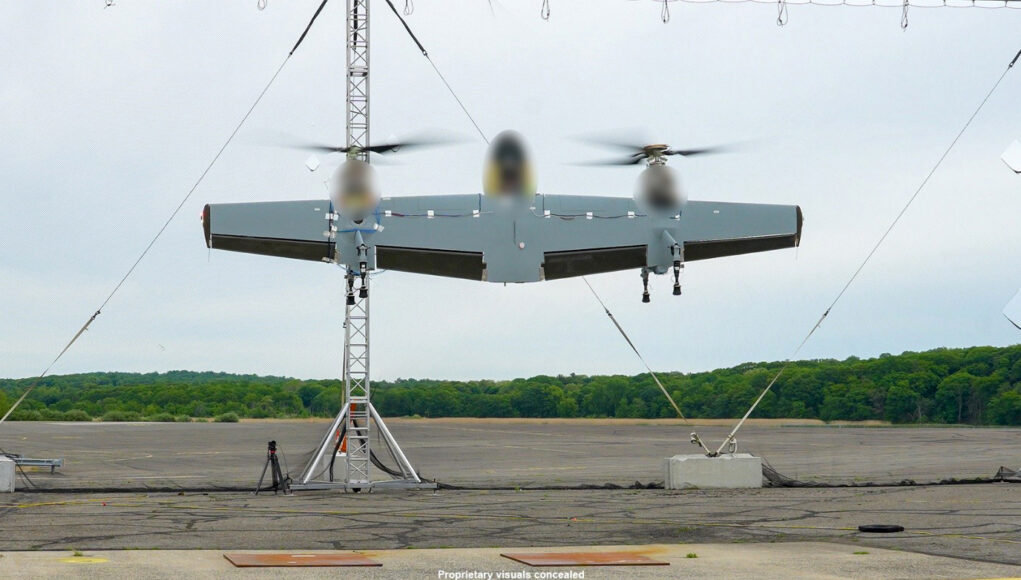 Three-rotor unmanned aircraft mounted on a test stand with supporting cables on a runway and a tree line in the background.