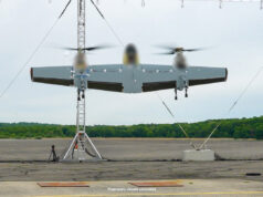 Three-rotor unmanned aircraft mounted on a test stand with supporting cables on a runway and a tree line in the background.