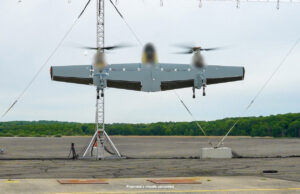 Three-rotor unmanned aircraft mounted on a test stand with supporting cables on a runway and a tree line in the background.