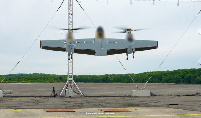 Three-rotor unmanned aircraft mounted on a test stand with supporting cables on a runway and a tree line in the background.