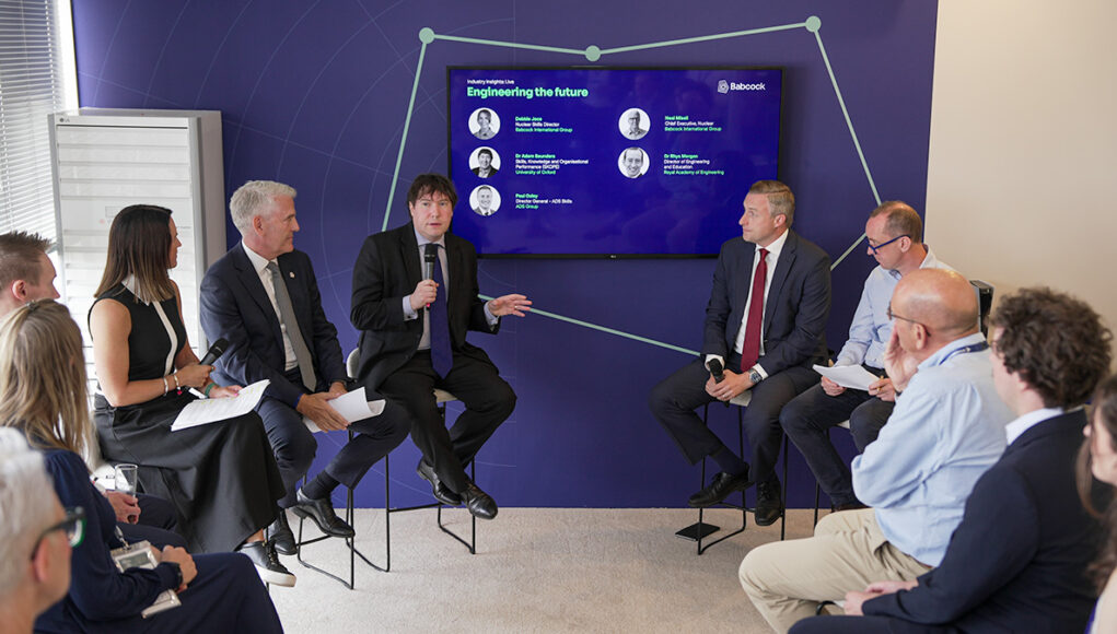 Panel discussion with six professionals in a blue-walled conference room, microphones in hand, a large screen behind shows 'Engineering the future'.