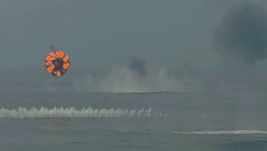 Duncan Circular fireball ring rising over the ocean from a distant explosion; smoke plumes in the sky above choppy water