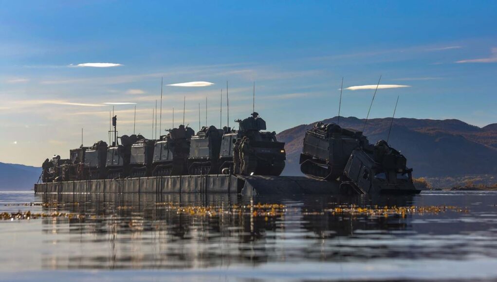 FLEET-20251001-ZI0059-002 Armored military vehicles loaded on a flat-bottom landing craft afloat on calm water with hills in the distance.