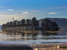 Armored military vehicles loaded on a flat-bottom landing craft afloat on calm water with hills in the distance.