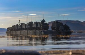 Armored military vehicles loaded on a flat-bottom landing craft afloat on calm water with hills in the distance.