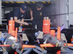 Two emergency responders in navy uniforms inspect equipment inside a transport vehicle, with orange safety posts in the foreground.