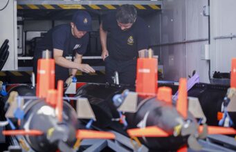 Minehunting teams embark warships ahead of global tasks Two emergency responders in navy uniforms inspect equipment inside a transport vehicle, with orange safety posts in the foreground.