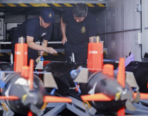 Minehunting teams embark warships ahead of global tasks Two emergency responders in navy uniforms inspect equipment inside a transport vehicle, with orange safety posts in the foreground.