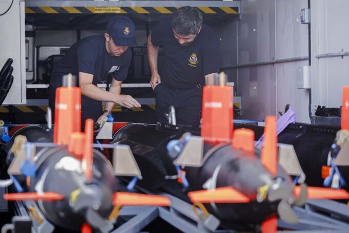 Minehunting teams embark warships ahead of global tasks Two emergency responders in navy uniforms inspect equipment inside a transport vehicle, with orange safety posts in the foreground.