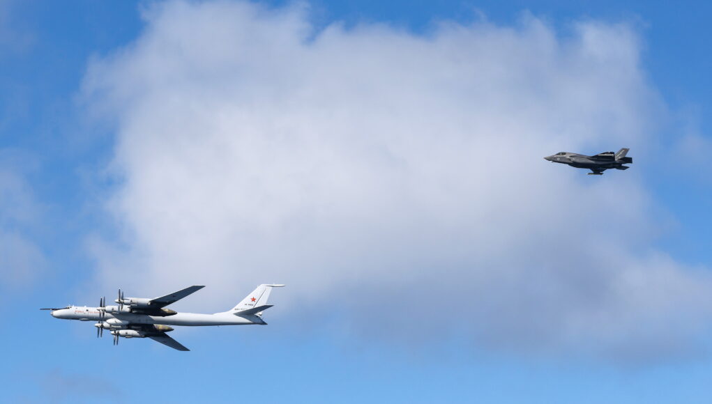 Royal Navy F-35B fighters of HMS Prince of Wales intercept a Russian military aircraft Two military aircraft in formation: a white four-engine propeller bomber on the left and a dark jet on the right against a blue sky with clouds.