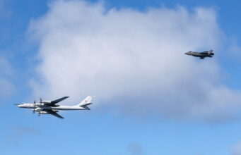 Two military aircraft in formation: a white four-engine propeller bomber on the left and a dark jet on the right against a blue sky with clouds.