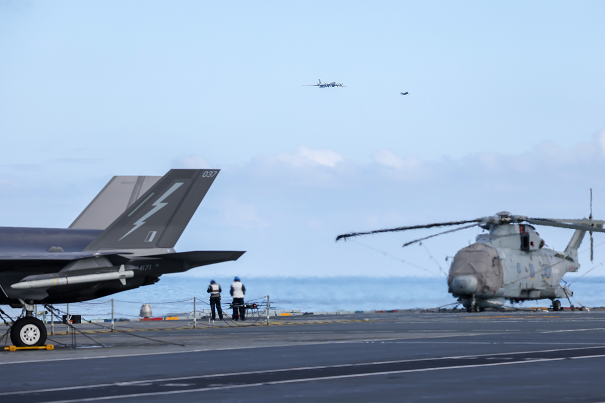 Carrier flight deck with a gray fighter jet on the left, a helicopter on the right, and crew near the edge against the ocean background.