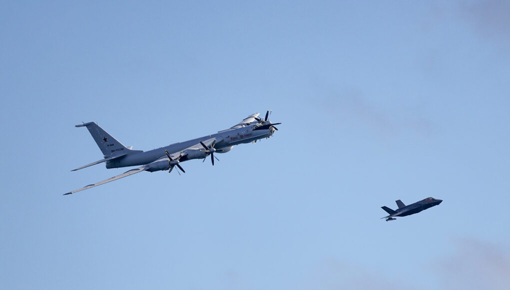 FLEET-20260702-XN0425-014 Large four-engine turboprop bomber in formation with a smaller fighter jet against a clear blue sky, mid-flight left to right.