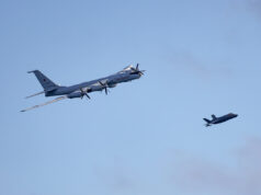 Large four-engine turboprop bomber in formation with a smaller fighter jet against a clear blue sky, mid-flight left to right.