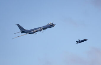 Russian Bear buzzes British aircraft carrier in Arctic Large four-engine turboprop bomber in formation with a smaller fighter jet against a clear blue sky, mid-flight left to right.