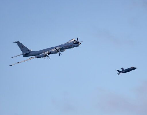 Large four-engine turboprop bomber in formation with a smaller fighter jet against a clear blue sky, mid-flight left to right.