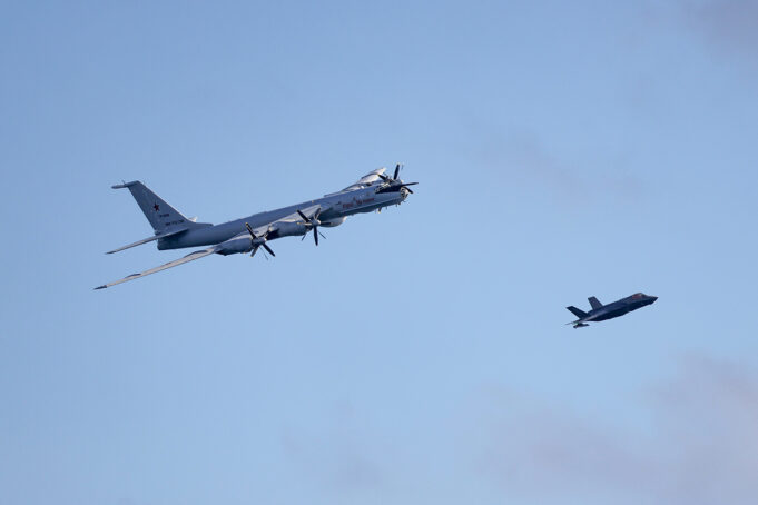 Russian Bear buzzes British aircraft carrier in Arctic Large four-engine turboprop bomber in formation with a smaller fighter jet against a clear blue sky, mid-flight left to right.