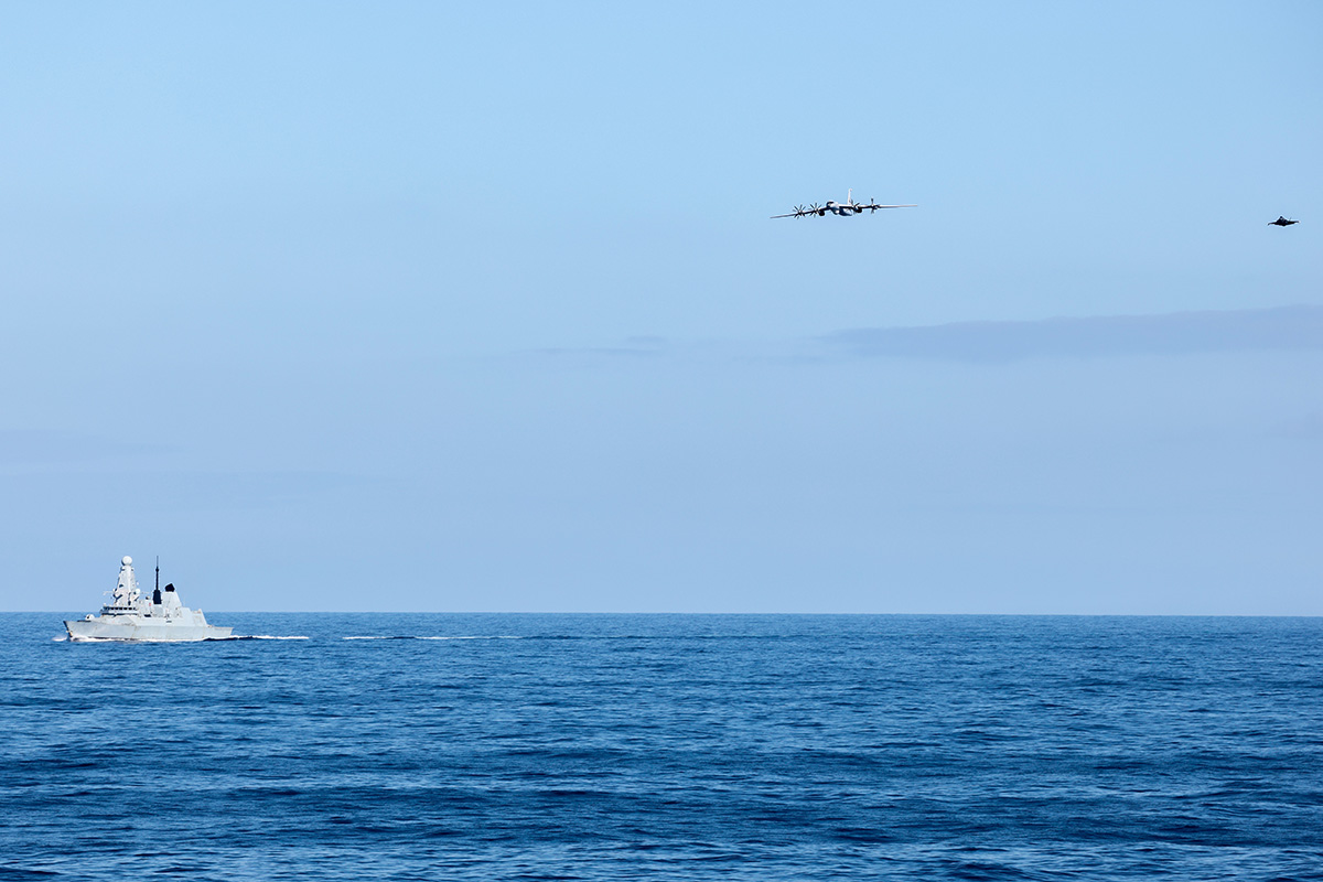 Gray military ship sails on calm blue sea with two aircraft flying overhead in formation against a clear sky at sea level.