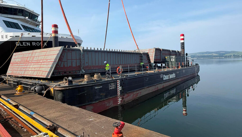 FMPG x BAE July 2026 2 Large barge with red steel panels and workers at a dock, unloading near a white passenger ferry ahead of calm water.