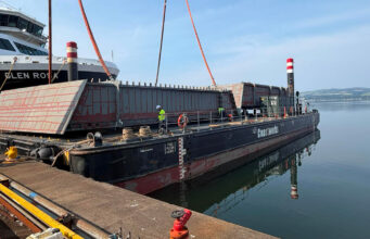 Large barge with red steel panels and workers at a dock, unloading near a white passenger ferry ahead of calm water.