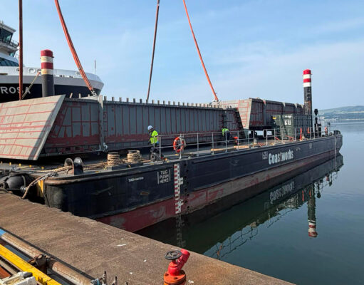 Large barge with red steel panels and workers at a dock, unloading near a white passenger ferry ahead of calm water.