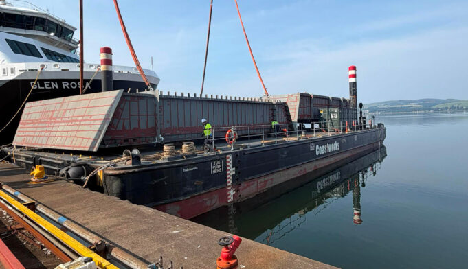 Ferguson Marine delivers Type 26 blocks to BAE on Clyde Large barge with red steel panels and workers at a dock, unloading near a white passenger ferry ahead of calm water.