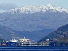Snowy mountains rise behind a harbor with ships moored along the quay, calm blue water in the foreground.