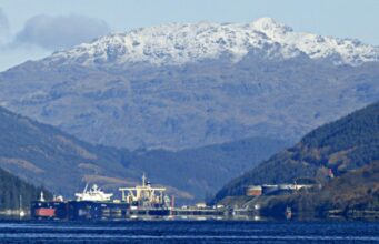 Snowy mountains rise behind a harbor with ships moored along the quay, calm blue water in the foreground.