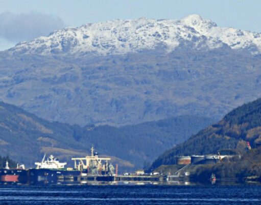 MoD buys Finnart oil terminal to boost Navy fuel reserves Snowy mountains rise behind a harbor with ships moored along the quay, calm blue water in the foreground.