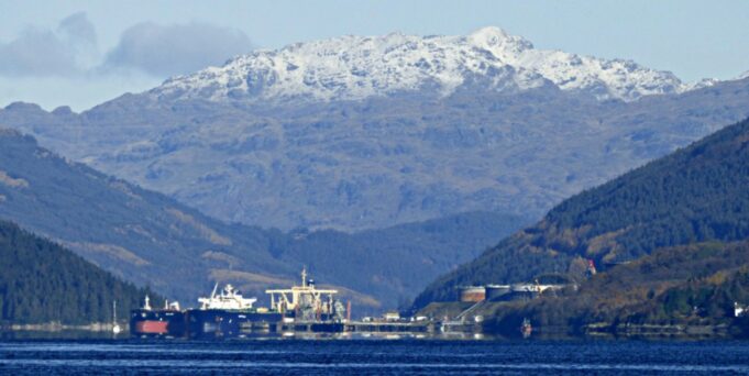 MoD buys Finnart oil terminal to boost Navy fuel reserves Snowy mountains rise behind a harbor with ships moored along the quay, calm blue water in the foreground.