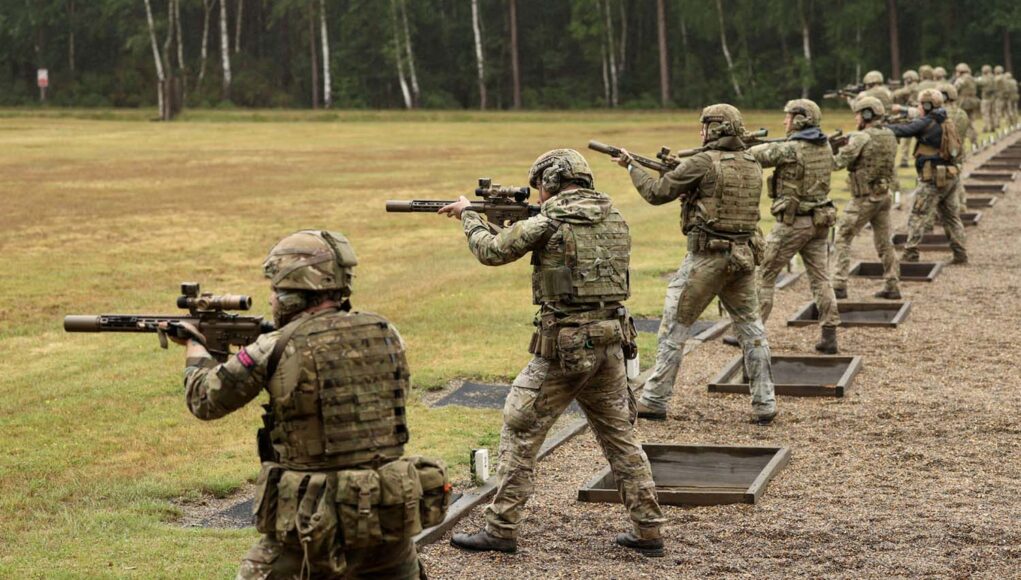 Formidable firepower - 14 shooters take aim standing up next to their foxholes Soldiers in camouflage gear firing rifles at an outdoor firing range, lined up in a row with tactical vests and helmets.