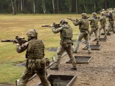 Soldiers in camouflage gear firing rifles at an outdoor firing range, lined up in a row with tactical vests and helmets.
