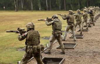 Soldiers in camouflage gear firing rifles at an outdoor firing range, lined up in a row with tactical vests and helmets.