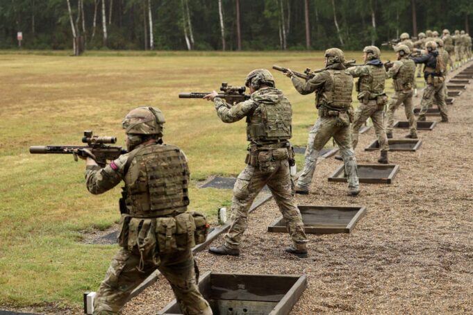 Soldiers in camouflage gear firing rifles at an outdoor firing range, lined up in a row with tactical vests and helmets.