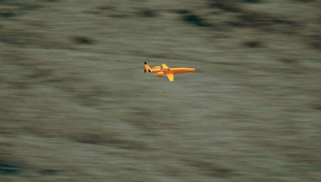 Greenjets LCADE2 Small orange propeller airplane in flight over a blurred grassy landscape, viewed from the side.