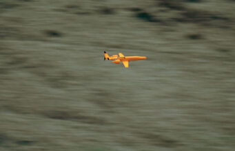 UK awards Europe’s first low-cost drone interceptor deals Small orange propeller airplane in flight over a blurred grassy landscape, viewed from the side.