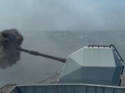 Military vessel at sea fires its main gun, ejecting a dense black smoke plume from the muzzle.