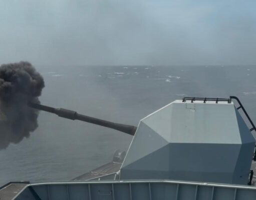 Military vessel at sea fires its main gun, ejecting a dense black smoke plume from the muzzle.