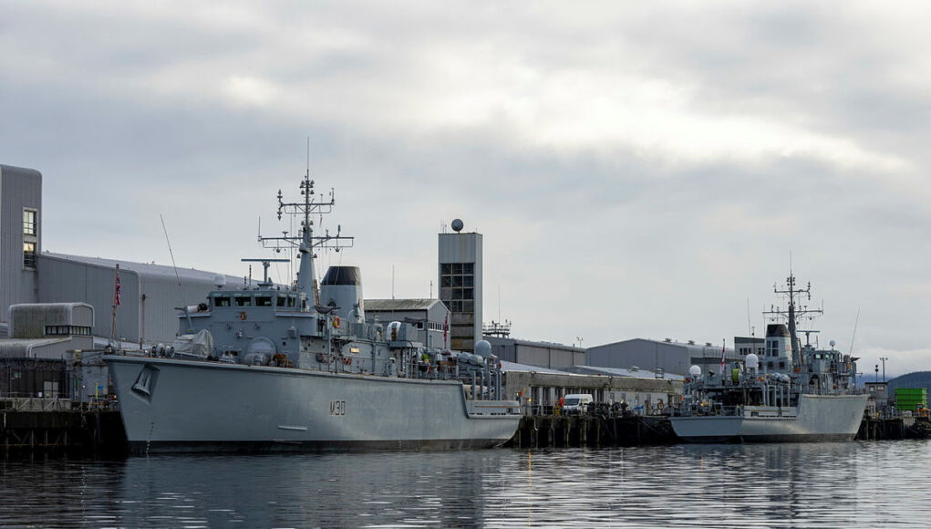 HUNT Two large grey military ships docked at a pier on a cloudy day, reflecting in calm water.