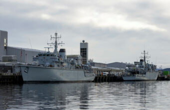Royal Navy’s last Hunt-class minehunter to go by 2031 Two large grey military ships docked at a pier on a cloudy day, reflecting in calm water.