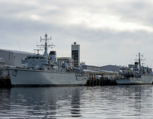 Two large grey military ships docked at a pier on a cloudy day, reflecting in calm water.