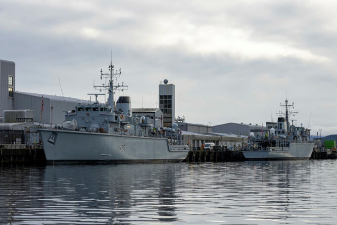 Royal Navy’s last Hunt-class minehunter to go by 2031 Two large grey military ships docked at a pier on a cloudy day, reflecting in calm water.
