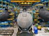 Inside a shipyard with three large submarine hulls being assembled, surrounded by scaffolding, yellow cranes, and industrial equipment.