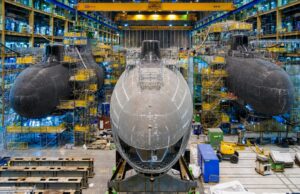 Inside a shipyard with three large submarine hulls being assembled, surrounded by scaffolding, yellow cranes, and industrial equipment.