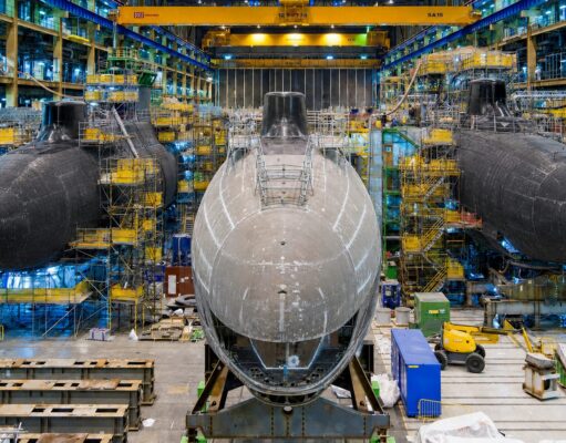 Britain to start building new nuclear attack subs next year Inside a shipyard with three large submarine hulls being assembled, surrounded by scaffolding, yellow cranes, and industrial equipment.