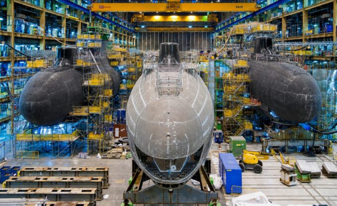 Britain to start building new nuclear attack subs next year Inside a shipyard with three large submarine hulls being assembled, surrounded by scaffolding, yellow cranes, and industrial equipment.